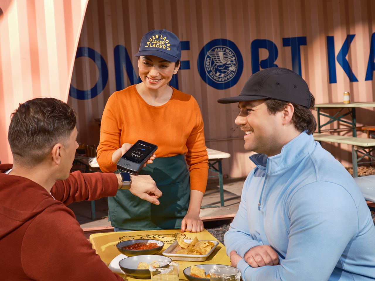 About Friends using smartwatch for contactless payment at an outdoor cafe, assisted by a smiling waitress.